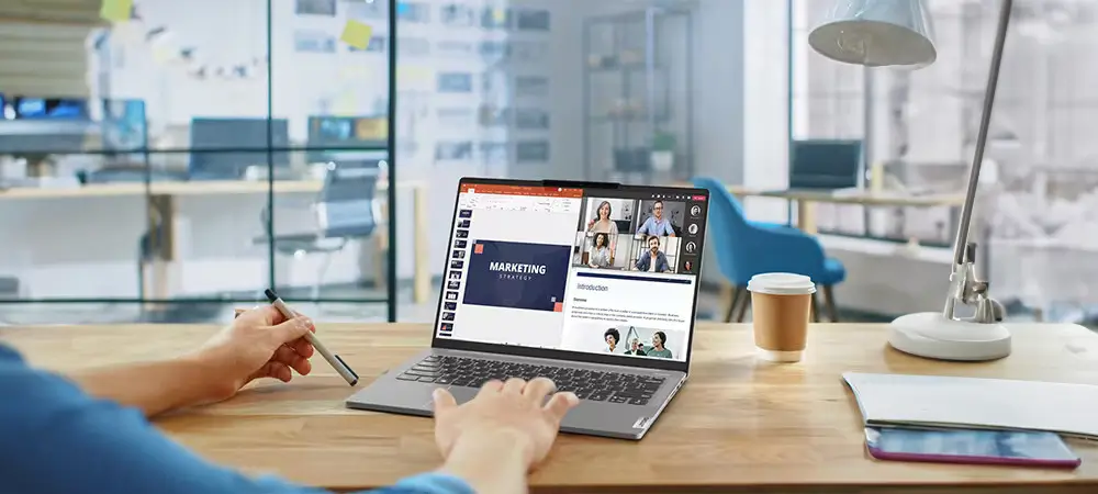 אדם יושב ליד שולחן עץ עם Lenovo IdeaPad Pro 5i מציג שיחת וידאו על חצי אחד של המסך ושקופית מצגת שכותרתה 